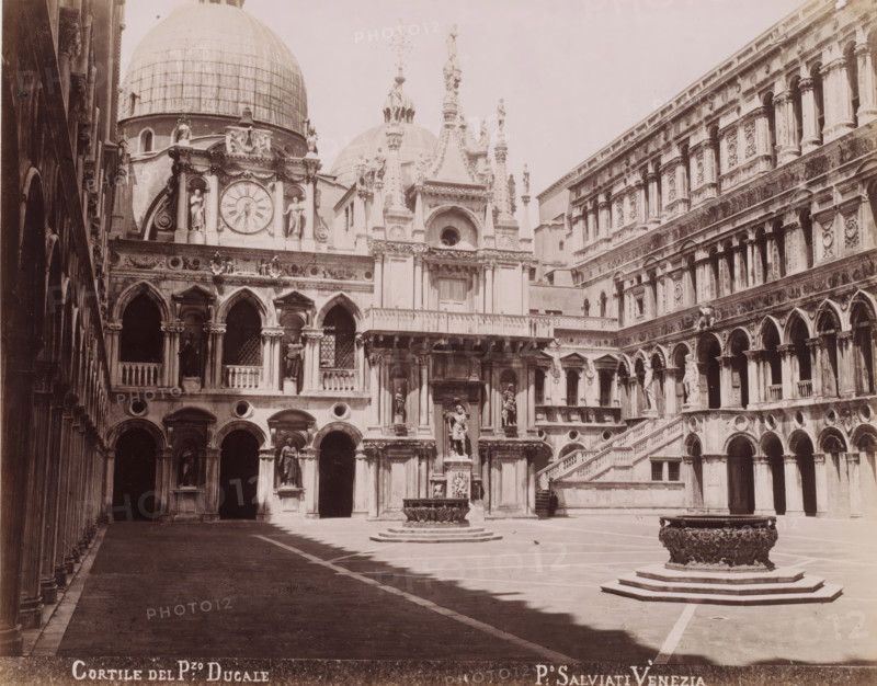 Cortile of the Doge's Palace in Venice, Cortile del P:zo Ducale, photograph, photographer: Paolo Salviati, Venice, 1860 - 1870, Paolo Salviati, photographer, pasteboard, paper, albumen print, albumendruk, height 196 mm x width 248 mm, height 275 mm x width 364 mm, palace, inner court of palace or 'hôtel', Doge's Palace, A large stone courtyard framed by ornate multi-story buildings with arched colonnades and carved facades fills the scene, featuring a central ornate wellhead on a raised stepped base and a second similar wellhead closer to the foreground, a wide staircase with balustrade rising along the right building, numerous sculpted statues set into niches and atop columns, decorative relief panels and circular windows, a clock face set within a richly ornamented section beneath domed roofs, shadow patterns across the paved ground, and a person standing near an entrance beneath the staircase.