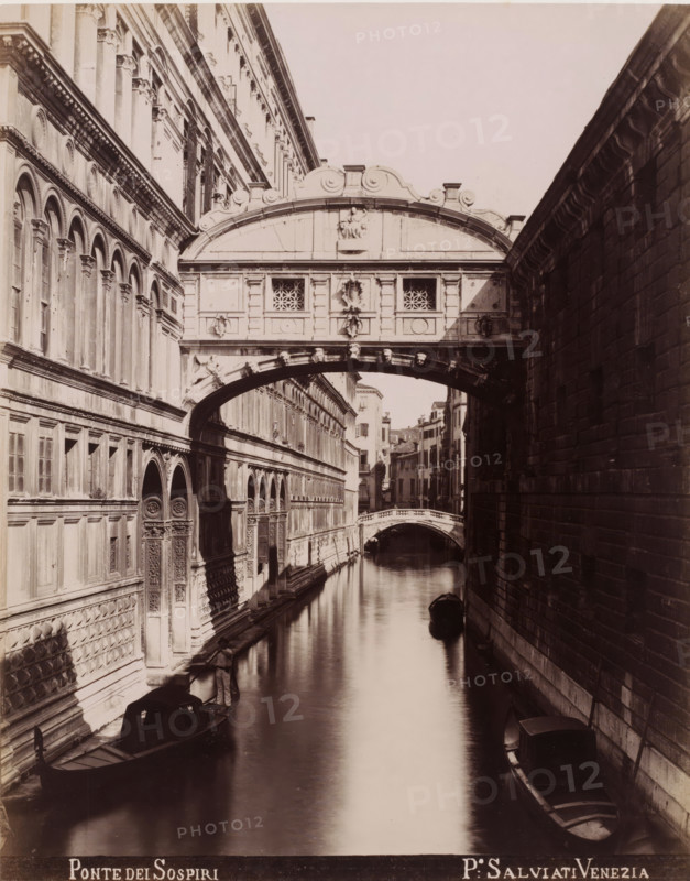 Ponte dei Sospiri, Bridge of Sighs in Venice, photograph, inscription: 'Venetie', photographer: Paolo Salviati, Venice, 1860 - 1870, pasteboard, paper, albumen print, albumendruk, height 251 mm x width 198 mm, height 365 mm x width 276 mm, bridge in city across river, canal, A narrow canal bordered by tall stone buildings with ornate facades and arched windows runs into the distance beneath a decorated enclosed bridge spanning the water, the bridge featuring carved panels and small lattice windows and sitting above a curved stone arch; gondolas float on the calm reflective water with one positioned near the foreground and another further back, and a person stands in a gondola along the left edge near a building entrance; the right building shows a textured masonry wall in shadow while the left building displays rows of arches, pilasters, and carved doorways, and a smaller arched pedestrian bridge is visible farther down the canal.