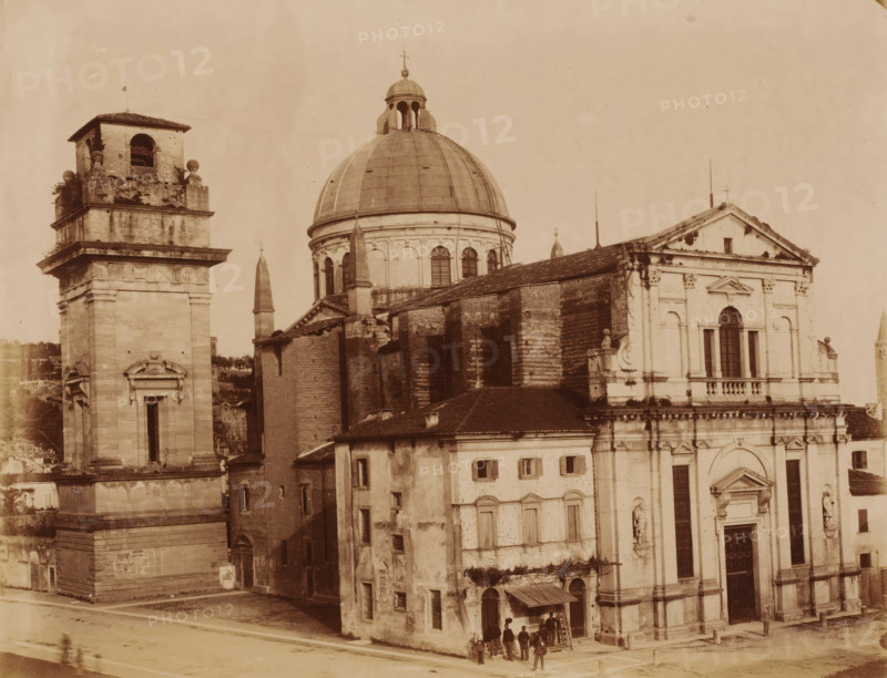 Exterior of the San Giorgio in Braida in Verona, Italy, photograph, inscription: 'Verona.', number: '13', Verona, 1851 - 1900, photographer, pasteboard, paper, albumen print, albumendruk, height 303 mm x width 405 mm, church (exterior), San Giorgio in Braida, A sepia-toned photograph showing a large classical church complex with a prominent domed roof and adjacent bell tower, stone façades with decorative cornices and pilasters, arched windows and sculpted niches, and a tiled sloping roof section with visible wear; the bell tower has an open belfry and ornamental spheres at its corners, while the church façade features a triangular pediment and a large arched window above a main entrance with heavy doors; several small rectangular windows and weathered brick and plaster walls are visible on the adjoining buildings, and a small group of people stands near a doorway under a simple awning at street level.