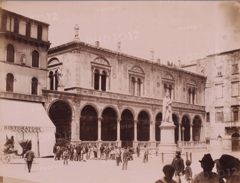 Statue of Dante on the Piazza dei Signori in Verona, Italy, photograph, number: '13', inscription: 'Verona.', Verona, after sculpture by Ugo Zannoni, 1851 - 1900, Ugo Zannoni, photographer, after sculpture by, pasteboard, paper, albumen print, height 305 mm x width 405 mm, Person, Dante Alighieri, monument, statue, square, circus, Piazza dei Signori, A wide facade of an ornate historic building with arched colonnades and decorative panels stretches across the image, topped by a row of statues standing on the roofline; a freestanding statue on a pedestal stands in the foreground to the right of center; groups of people and children gather and walk in the open plaza in front of the building, some near a horse-drawn carriage and others near an awninged storefront at the left edge; shuttered windows and architectural ornamentation are visible on adjacent buildings framing the scene.