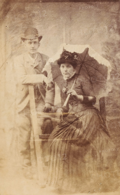 Portrait of an unknown woman with parasol and an unknown man, photograph, carte-de-visite, 1851 - 1900, photographer, inorganic material, glass, paper, albumen print, with glass cover plate, height 104 mm x width 62 mm, height 109 mm x width 69 mm x depth 4 mm, adult woman, adult man, anonymous historical persons depicted in a double portrait, parasol, sunshade, A sepia photograph of a man and a woman posed together; the man stands to one side wearing a jacket and a hat with one arm resting on a wooden prop, while the woman sits holding a parasol above her shoulder and wears a dark dress with a high collar, gloves, and a decorated hat. The woman's skirt has visible vertical pleats and texture, and the parasol has a scalloped edge. Both faces are visible with neutral expressions. The background is plain with a mottled studio backdrop, and the image shows signs of age such as fading and small spots.