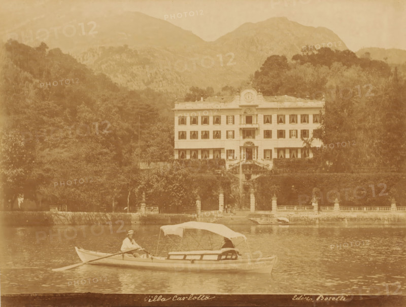 Villa Carlotta, Villa Carlotta on Lake Como, Tremezzo, Italy, photograph, publisher: Bosetti, 1851 - 1900, photographer, Lombardy, pasteboard, paper, albumen print, height 348 mm x width 455 mm, villa, lake, rowing facing backward, pulling, Lake Como, A sepia-toned photograph shows a large multi-story villa with symmetrical rows of windows and a central entrance flanked by stairways, set behind a tree-lined terrace and balustrade; in front of the villa a body of water reflects the scene and a small rowboat with a partially covered canopy floats on the water, carrying two people, one seated at the oars wearing a hat, with wooded hills and mountains rising in the background.
