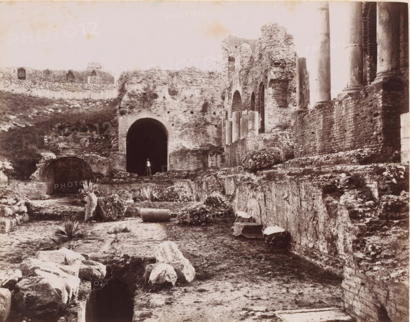 Ruin of the Teatro Greco in Taormina, Italy, Teatro Greco, Taormina, photograph, inscription: 'RBK', number: '8663', number: '21', photographer: Giorgio Sommer, 1857 - 1914, Sicily, pasteboard, paper, albumen print, height 320 mm x width 369 mm, ruin of a building, architecture, excavation, archaeology, Greek theater, A wide sepia photograph of ancient ruins showing crumbling brick and stone walls, arches, and columns extending across the scene, with a large dark arched entrance near the center and a person standing in front of it, scattered broken stone blocks and fragments in the foreground, low vegetation and a few agave-like plants among the ruins, weathered cylindrical column pieces lying on the ground, and tall intact columns rising at the right edge against a pale sky.