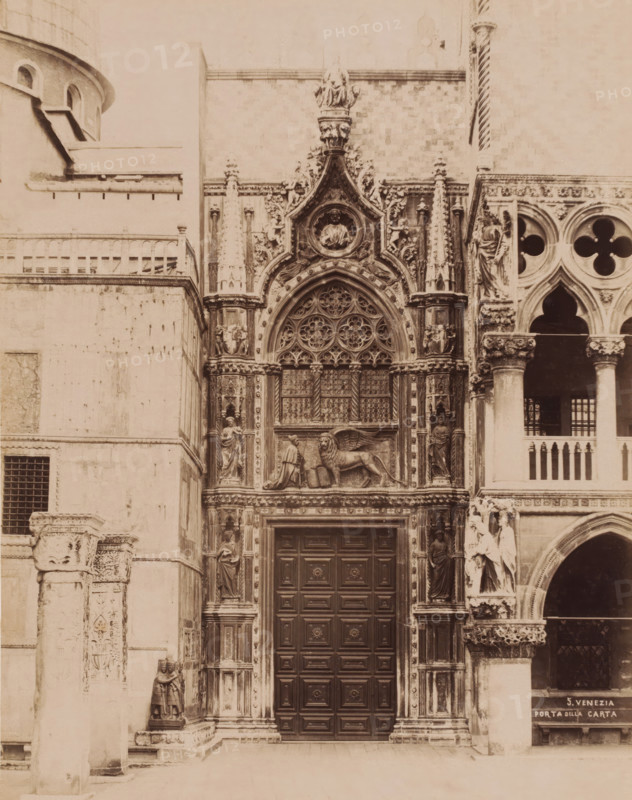 Porta della Carta in the Basilica of San Marco in Venice, Italy, Porta della Carta, Venezia, photograph, number: '5.', Venice, 1851 - 1900, photographer, pasteboard, paper, albumen print, albumendruk, height 369 mm x width 322 mm, parts of church exterior and annexes: portal, Florence, Tuscany, San Marco, A sepia-toned photograph of an ornate stone building facade showing a large wooden door with raised rectangular panels set within a richly carved archway decorated with tracery and circular floral motifs; flanking the doorway are sculpted columns and niches containing standing human figures, and above the arch is a relief of a winged lion next to a robed kneeling figure beneath a circular medallion containing a sculpted bust, all set among elaborate foliage and architectural ornament; to the right a section of arcade with rounded arches and quatrefoil openings is visible, with additional sculpted figures and ornate capitals, while to the left plain stone walls and a small barred windo...