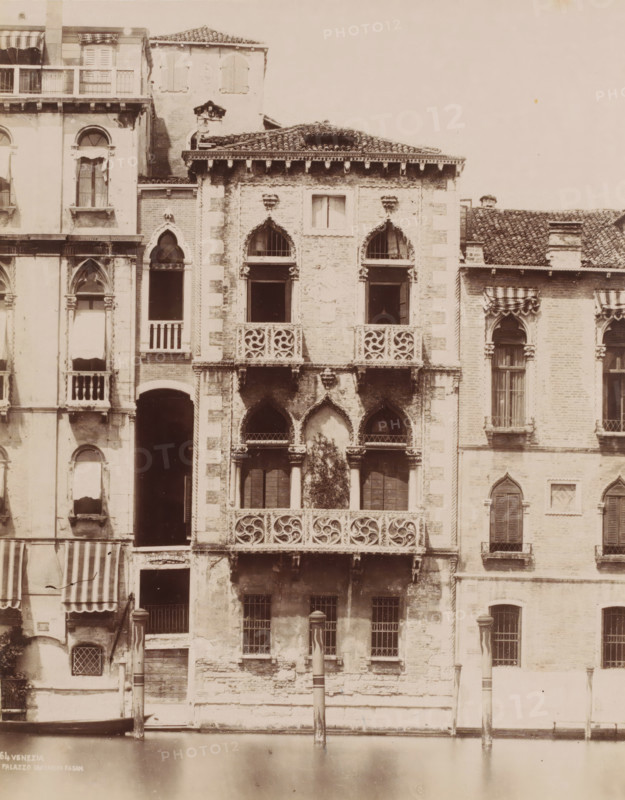Palazzo Contarini Fasan, Façade of the Palazzo Contarini Fasan in Venice, Italy, Venezia, photograph, number: '64', Venice, 1851 - 1900, photographer, pasteboard, paper, albumen print, albumendruk, height 368 mm x width 318 mm, canals, waters (in city), facade (of house or building), A tall façade of connected old buildings rises from a calm canal, showing weathered stone and brick walls with decorative balconies and arched windows; ornate carved balustrades and pointed arch openings appear on the central section, with two small columns flanking a darker arched entrance near the waterline and a pair of striped poles standing in the canal; adjacent sections display shuttered and open windows with fabric awnings, stone cornices and tiled roofs, and areas of peeling plaster and exposed brickwork.