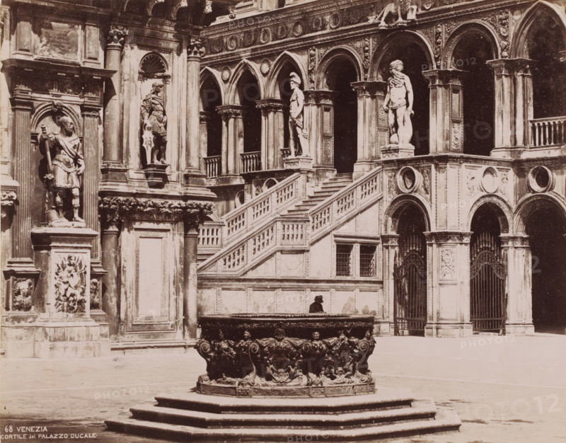 Courtyard of the Doge's Palace in Venice, Italy, Cortile del palazzo Ducale, Venezia, photograph, number: '68', Venice, 1851 - 1900, photographer, pasteboard, paper, albumen print, albumendruk, height 319 mm x width 368 mm, inner court of palace or 'hôtel', Doge's Palace, A stone courtyard scene showing a large ornate circular fountain basin on a stepped platform in the foreground, the basin decorated with sculpted faces and ornamental reliefs. Behind it a grand exterior wall of a palatial building with carved columns, arched openings, decorative medallions, and balustraded stairs leading up to a terrace. Several life-sized stone statues stand in niches and on pedestals along the facade, including figures in classical poses and draped garments, and one bearded figure. There are wrought iron gates set into arched doorways and carved panels and reliefs filling many of the architectural surfaces. A solitary figure sits on the edge of a raised platform near the base of the staircase. The i...