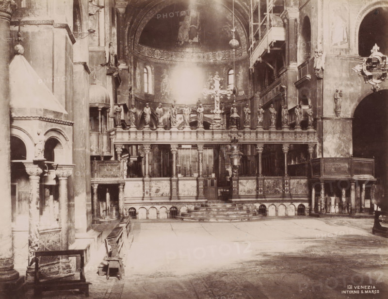 Interno S. Marco, Interior of San Marco in Venice, Italy, Venezia, photograph, number: '131', Venice, 1851 - 1900, photographer, pasteboard, paper, albumen print, albumendruk, height 320 mm x width 365 mm, interior of church, Florence, Tuscany, San Marco, A sepia-toned interior view of a large ornate space dominated by a raised altar area with a balustrade topped by a row of standing statues and a central decorative cross, framed by columns and arches; behind the altar a semicircular apse with painted figures and small arched windows rises beneath an embellished arch, while scaffolding and a suspended lamp are visible to one side; the foreground shows a wide open floor with worn stone paving, early wooden benches along the left, carved pillars and arched openings, and various carved reliefs and architectural details on the walls and cornices throughout the scene.