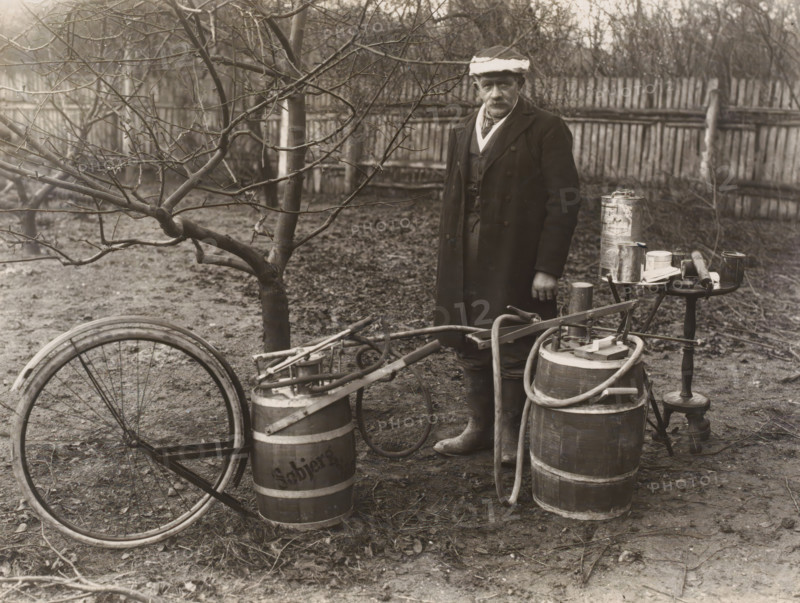 Man by a self-built pesticide spraying system for fruit trees, possibly in Denmark, photograph, Denmark, 1900 - 1930, photographer, paper, baryta paper, gelatin silver print, developing gelatin silver print, height 171 mm x width 228 mm, adult man, fruit-tree culture, fruit harvest, A person wearing a long dark overcoat, layered clothing, boots, and a hat stands near wooden barrels and a small round table holding metal cans and a few tools, with hoses and a hand-operated pump attached to the barrels; a bare-branched tree and a wooden fence form the background, and a bicycle wheel and frame lie on the ground among scattered leaves and dirt.
