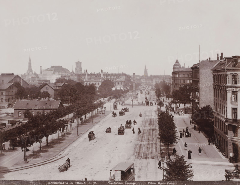 View of the Vesterbrogade in Copenhagen with the Hovedbanegård on the left and the Frihedsstøtten, Vesterbros Passage, Kjøbenhavn og Omegn in the middle, photograph, number: 'Nr. 27.', Copenhagen (city), publisher: Vilhelm Trydes, Denmark, 1880 - 1910, photographer, paper, pasteboard, baryta paper, daylight collodion silver print, height 217 mm x width 283 mm, height 350 mm x width 455 mm, monument, statue, street, Københavns Hovedbanegård, Liberty Memorial, Vesterbrogade, A wide urban street scene seen from above with parallel tracks running down the center and a long avenue lined with trees and lampposts, horse-drawn carriages and early motor vehicles moving along the road and pedestrians walking on the pavements, a tall column monument near the middle of the street, multi-story buildings with detailed facades and rooftop chimneys on the right side, rows of trees and lower buildings on the left, distant domed and spired architectural structures on the skyline, and people in period cl...