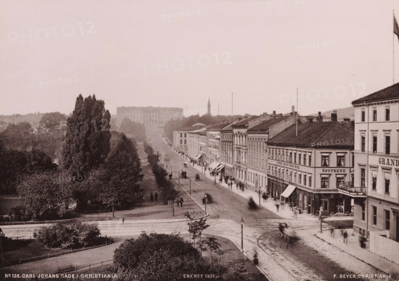 Carl Johans Gade i Christiania, View of the Karl Johans gate in Oslo, photomechanical print, copyright notice: 'ENERET 1891.', number: 'No 138.', Oslo, publisher: F. Beyer, Bergen, cliché maker: anonymous, 1891, photographer, paper, collotype, photoprint, height 151 mm x width 212 mm, street, Christiania, A wide sepia-toned street scene with a tree-lined promenade on the left and a row of multi-story buildings with awnings and shopfronts on the right, a paved road running between them with visible tram rails and blurred figures and vehicles in motion, lamp posts along the roadway, dense clusters of trees and shrubs in the foreground and midground, a large classical building set back among trees in the distance, a tall church spire rising behind the treeline, and a flagpole with a flag at the far right atop one of the corner buildings.