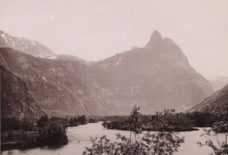 View of the Romsdalshornet in Norway, Romsdalshorn og vængetinderne, photomechanical print, number: 'No 219.', copyright notice: 'ENERET 1891.', Norway, publisher: F. Beyer, Bergen, cliché maker: anonymous, 1891, photographer, paper, collotype, photoprint, height 154 mm x width 212 mm, mountains, Romsdalshornet, A wide landscape showing a river flowing toward the foreground with low trees and shrubs along its banks, a broad flat plain beyond the near water, steep rocky mountain slopes rising on both sides, a prominent jagged peak on the right with patches of snow on its upper ridges, additional serrated snowy ridges visible on the left, and a pale sky above.