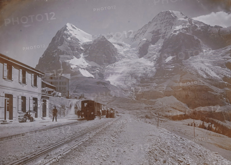 Wengernalp with Eiger and Mönch, View of a railway track in Wengen at the foot of the Eiger and the Mönch in the Bernese Alps, Hotel Jungfrau on the left, photograph, number: '3664.', Wengen, 1890 - 1930, photographer, paper, baryta paper, daylight gelatin silver print, height 164 mm x width 220 mm, railway, train, mountains, A mountainous landscape fills the scene with snow-covered peaks and exposed rock faces rising in the background, and a large glacier flowing down between steep cliffs. In the foreground a railway track runs toward the mountains with a small train stopped on the line and a group of people gathered near the cars. To the left, a multi-story building with shuttered windows lines the platform where a few individuals sit on a bench and another stands nearby; another building sits slightly back on a slope. The ground near the tracks is gravelly and uneven, and the right side of the image shows sloping terrain with scattered rocks and sparse vegetation.