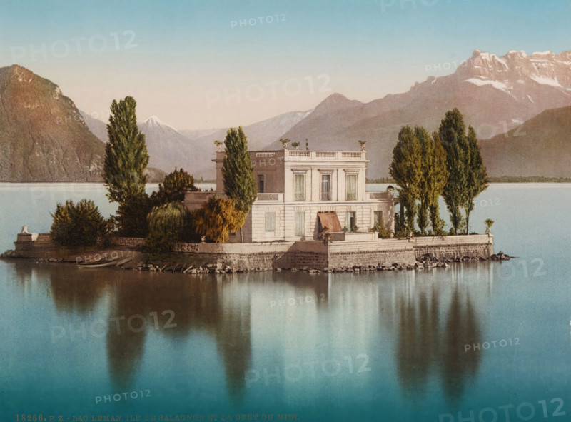 View of Île de Salagnon in Lake Geneva, in the background the Dents du Midi, lac Léman, Ile de Salagnon et la Dent du Midi, photomechanical print, number: '18266.', Lake Geneva, cliché maker: Photochrom Zürich, Zürich (city), publisher: Photochrom Zurich, Zurich (city), 1889 - c. 1920, photographer, paper, photochrom, height 166 mm x width 224 mm, island, villa, lake, mountains, Alps, Dents du Midi, Île de Salagnon, A pale rectangular villa sits on a stone quay projecting into calm turquoise water, flanked by tall columnar trees and rounded shrubs that cast soft reflections on the water surface; the two-story building features shuttered windows, a small awning over a doorway, decorative balustrades along the roofline, and potted plants on the terrace; the surrounding shoreline shows rocky edges and a low stone wall, while distant mountains rise behind the villa, some with snow-capped peaks under a clear sky.