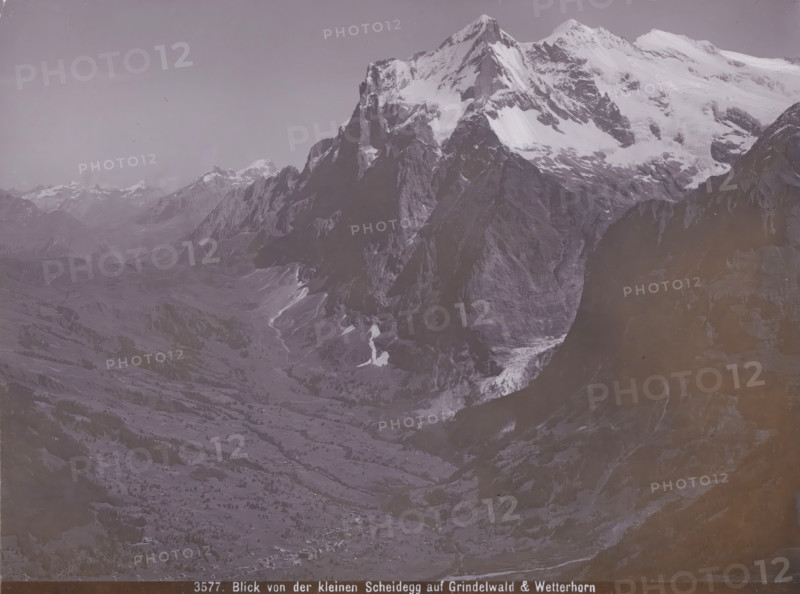 View of the Kleinen Scheidegg auf Grindelwald & Wetterhorn, View of the Grindelwald and the Wetterhorn from the Kleine Scheidegg, photograph, number: '3577.', Kleine Scheidegg, 1890 - 1930, photographer, paper, baryta paper, daylight gelatin silver print, height 164 mm x width 219 mm, mountains, Grindelwald, Wetterhorn, A wide mountainous landscape with a large snow-capped peak dominating the right side and rugged dark rock faces beneath the snow, tapering down into steep cliffs and a deep valley; patches of snow and ice sit on ledges and in gullies, and lower slopes show grassy and rocky terrain with scattered trees and small clusters of buildings visible near the valley floor; distant mountain ridges and peaks extend across the left and center background under a pale, cloudless sky.