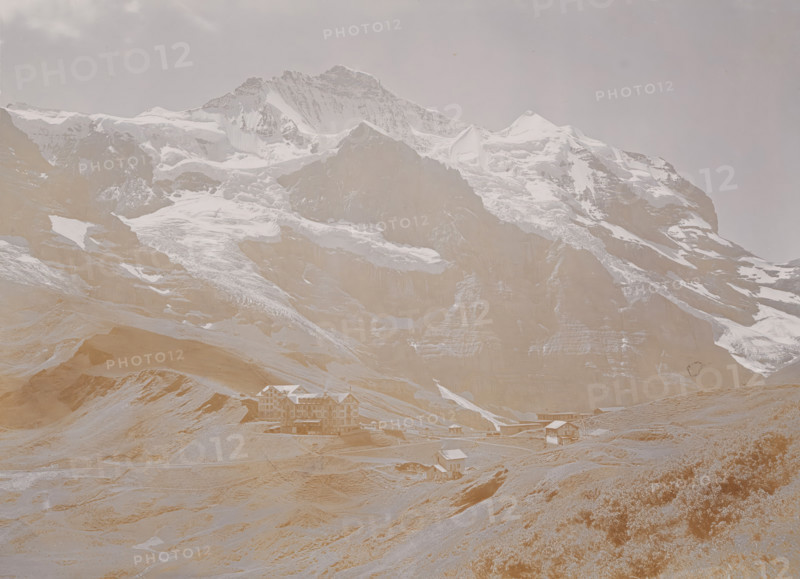 Kleine Scheidegg, View of the Kleine Scheidegg in the Bernese Alps, photograph, number: '3678.', Berner Alpen, 1880 - 1910, photographer, Alps, Bernese Alps, paper, baryta paper, daylight collodion silver print, height 165 mm x width 219 mm, mountains, A wide mountain scene showing a large rocky peak with snow and ice on its upper slopes and ridges under a pale sky, with steep cliffs and layered rock faces below; grassy and rocky foothills lead down from the mountain, where several multi-story chalet-style buildings and smaller wooden structures sit scattered along a winding road and plateau near the base of the slopes.