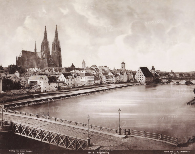 View of Regensburg, Germany, Regensburg, photomechanical print, number: 'Nr. 4.', cliché maker: Johann Baptist Obernetter, Munich, publisher: Oscar Krämer, Vienna, 1856 - 1887, photographer, Bavaria, Lower Austria, pasteboard, paper, collotype, photoprint, height 305 mm x width 372 mm, city-view in general, 'veduta', A riverside cityscape with a wide calm river in the foreground, a wooden bridge crossing the river with visible railings and lampposts, and rows of multi-story buildings with pitched roofs lining the riverbank; a large Gothic cathedral with two tall spires rises prominently behind the buildings, and several towers and a distinctive sloped-roof warehouse or granary appear among the rooftops; a stone arched bridge connects the far bank, and the sky above is filled with pale, soft clouds.