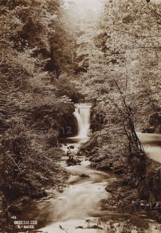 Waterfall with standing boy, photograph, date: '1903', inscription: '148., Gust. Salzer, B.-Baden.', photographer: Gustav Salzer, Baden-Baden, 1903, Baden-Württemberg, paper, baryta paper, daylight collodion silver print, height 168 mm x width 117 mm, waterfall, A sepia-toned scene of a forested ravine with a waterfall cascading into a stream that flows toward the foreground, surrounded by dense trees and undergrowth on both banks, rocky outcrops and boulders lining the watercourse, a lone person wearing a hat seated on a rock near the stream below the waterfall, and a narrow path visible along the right bank beside leaning and twisted tree trunks.