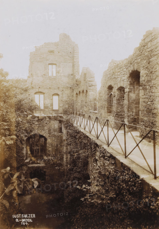 Ruin of Hohenbaden Castle, photograph, inscription: 'Gust. Salzer, B.-Baden., 154.', date: '1902', photographer: Gustav Salzer, Baden-Baden, 1902, Baden-Württemberg, paper, baryta paper, daylight collodion silver print, height 166 mm x width 116 mm, ruin of a dwelling, house, castle, A sepia-toned photograph showing the stone ruins of a building with a long narrow walkway edged by a metal railing running alongside a high stone wall with arched openings, a taller ruined facade with rectangular window openings at the far end, overgrown vegetation and ivy clinging to the lower walls and ground below, and a dark arched doorway or opening at the base partially obscured by foliage.