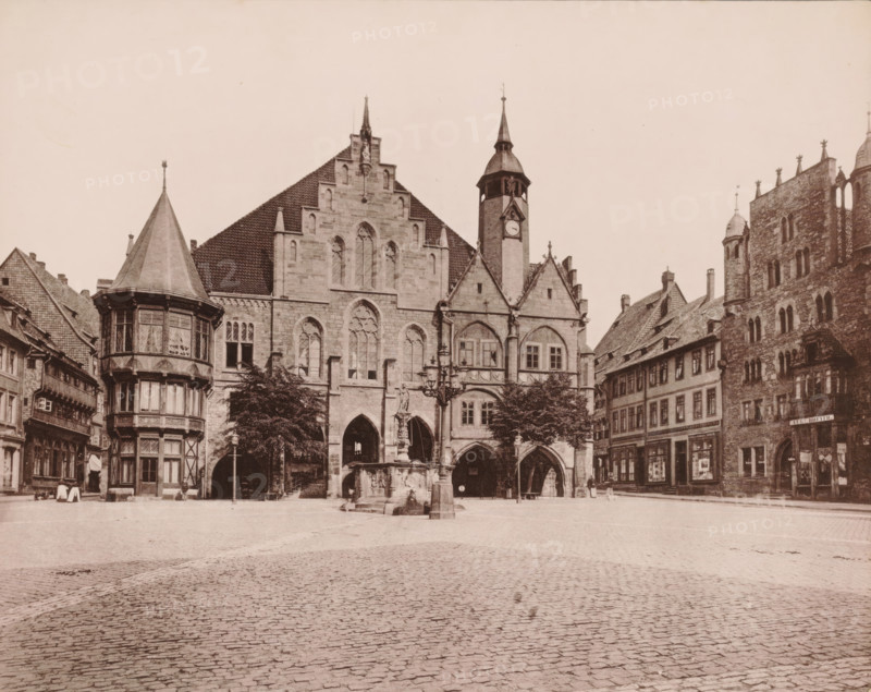 Hildesheim Town Hall, photomechanical print, inscription: 'Hildesheim', number: 'N.1.', photographer: E. Mertens & Cie (possibly), Hildesheim, cliché maker: anonymous, 1880 - 1900, Lower Saxony, paper, collotype, photoprint, height 228 mm x width 286 mm, square, circus, townhall, A wide cobblestone square fills the foreground leading to a row of ornate stone and timber buildings along the back edge. Prominent in the center is a large Gothic stone building with pointed arched openings at ground level, tall lancet windows above, stepped gables, and a clock tower with a spire rising to the right. In front of this building stands a sculpted fountain with a central column and a decorative lamp post nearby. To the left a multi-story bay with a conical roof and patterned window panes projects over the street, flanked by smaller timber-framed buildings. To the right of the central structure are steeply pitched tiled roofs and a tall stone building with narrow arched windows and decorative roof...