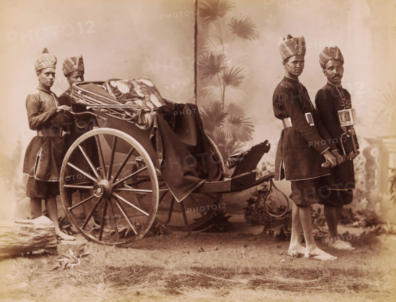 Studio portrait of four Indian men in uniform with a carriage, India, photograph, inscription: '£1', inscription: 'Asia', signature: 'SH Dagg', photographer: Sidney Herbert Dagg, British India, 1890 - 1910, pasteboard, paper, albumen print, height 213 mm x width 275 mm, height 227 mm x width 293 mm, clothes for official occasions, rickshaw, A sepia-toned photograph showing a large wooden two-wheeled carriage draped with patterned fabric and a dark cloth, flanked by four men in matching dark uniforms and tall patterned turbans; two men stand near the rear wheel holding the carriage frame while two stand at the front holding the shafts, one of them carrying a box-like lantern attached to the shaft, and all four are barefoot on a grassy surface with a painted backdrop of tall, leafy plants behind them.