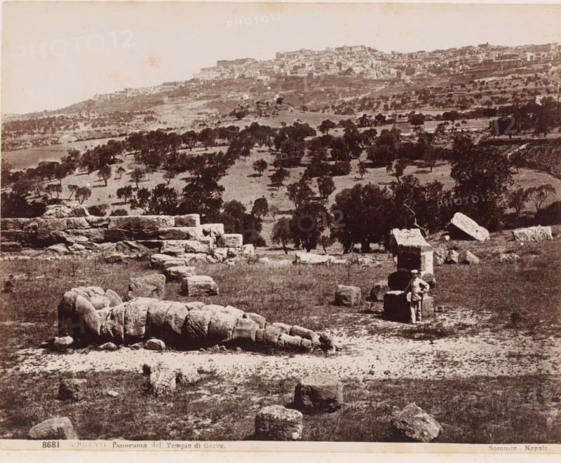 View of Agrigento from the Temple of Jupiter, Italy, Panorama dal Tempio di Giove, Girgenti, photograph, number: '8681', photographer: Giorgio Sommer, Agrigento, 1857 - 1914, Sicily, pasteboard, paper, albumen print, height 320 mm x width 369 mm, city-view in general, 'veduta', dale, valley, A sepia-toned landscape showing a hillside town in the distance above a gently sloping plain dotted with scattered trees and low vegetation, with large rough-cut stone blocks and fragments scattered across the foreground and a stepped stone structure extending across the middle ground, and a solitary person standing beside a rectangular stone block near the right side of the image.