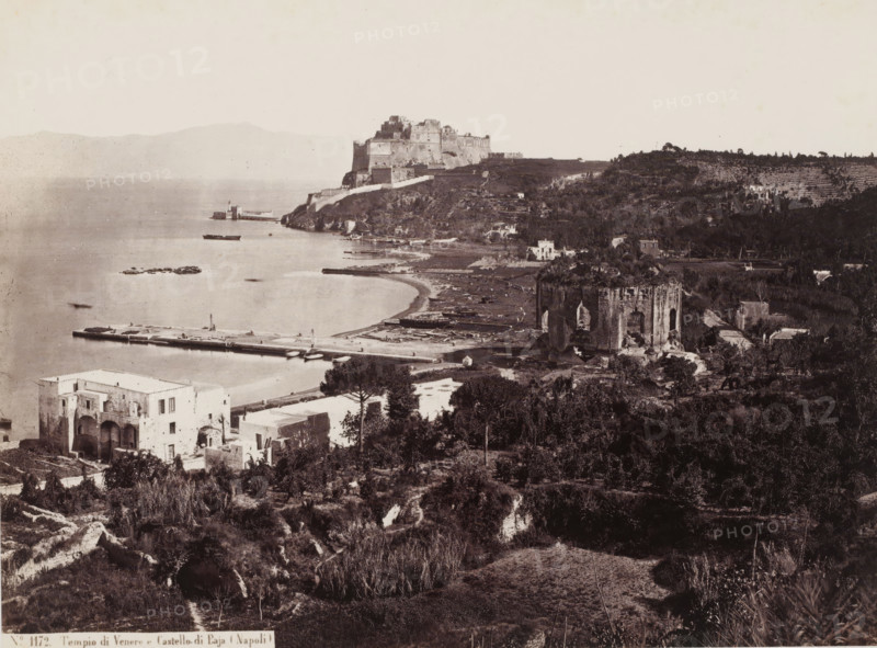 View of the Bay of Naples, Italy, Tempio di Venere e Castello di Baja Napoli, photograph, number: 'No. 1172.', address: 'GIORGIO SOMMER, STUDIO MONTE DI DIO 4., MAGAZZINO S. CATERINA 5., NAPOLI', photographer: Giorgio Sommer, Naples, 1857 - 1914, pasteboard, paper, albumen print, albumendruk, height 306 mm x width 375 mm, gelding of a building, architecture, gulf, bay, fortress, A wide coastal panorama showing a body of calm water with a long pier extending into it and small boats moored nearby, a curved shoreline with a narrow beach, a large fortress-like structure perched on a headland above the shore, terraces and dense vegetation covering hillsides, multiple stone and stucco buildings of varying sizes scattered among trees, a prominent ruined tower or cylindrical stone structure near the shoreline with visible openings and weathered surfaces, agricultural plots and rows of planted terraces on the distant hillside, and a foreground of shrubs, low trees, and uneven ground leading dow...