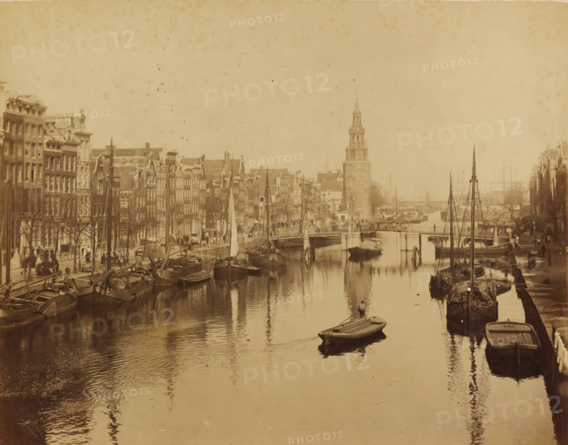 Oude Schans and Montelbaan Tower in Amsterdam, photograph, photographer: Albert Greiner, Amsterdam, 1861 - 1889, pasteboard, paper, albumen print, height 268 mm x width 342 mm, height 484 mm x width 654 mm, urban housing, canal, ships (in general), quay, Oudeschans, Montelbaan Tower, A sepia-toned city scene showing a canal lined with narrow multi-story buildings on both sides, with numerous moored sailboats and rowboats along the water edges and reflections on the canal surface, a small rowboat with a person rowing toward the foreground, a stone bridge crossing the canal further back, and a tall clock tower rising near the bridge with streets and people visible along the quay.