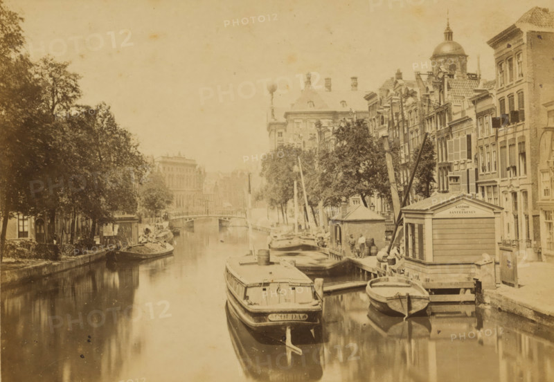 The Pipe Market, View of the Pipe Market in Amsterdam, Amsterdam, cabinet photograph, annotation: 'NZ Voorburgwal - pipe market, and old flower market, filled in 1884.', publisher: Andries Jager, 1860 - 1884, photographer, paper, pasteboard, albumen print, height 109 mm x width 167 mm, canals, waters (in city), A narrow waterway runs through the scene with several moored boats of varying sizes along the right bank and one large vessel prominent in the foreground, its bow reflected in the water; wooden docks, barrels, and small boathouses line the right edge of the canal. Tall, narrow multi-story buildings with shuttered windows and decorative facades rise behind the docks, interspersed with leafy trees whose canopies extend over parts of the canal. On the left bank a row of trees and low buildings create a shaded promenade with a small boat tied up at the edge. A bridge spans the canal in the mid distance beyond which larger stone buildings and architectural details are visible, and a ...