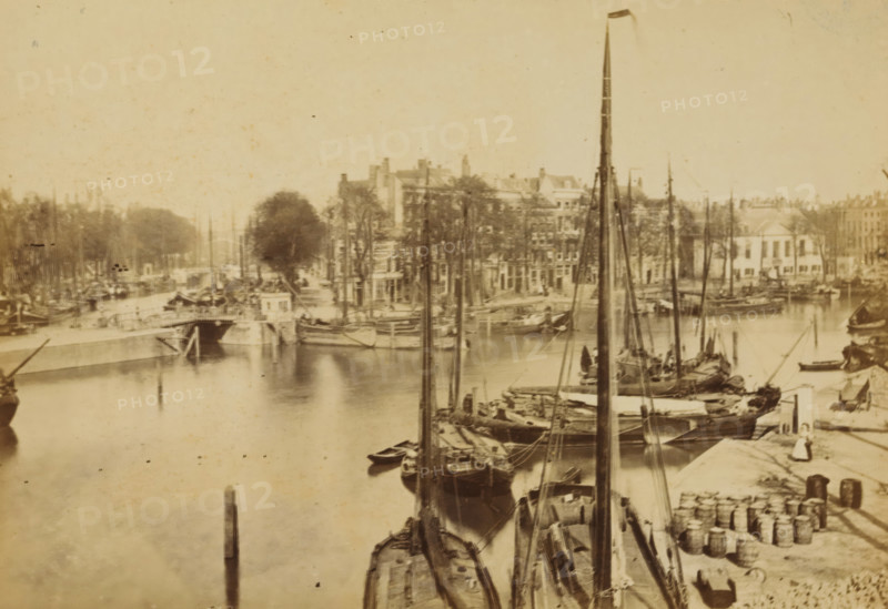 The Spanish Quay, View of the Spaansekade in Rotterdam, Rotterdam, cabinet photograph, annotation: '45, 5, 40, 12x: 15, 55', publisher: Andries Jager, Amsterdam, 1860 - 1890, photographer, paper, pasteboard, albumen print, height 108 mm x width 168 mm, canals, waters (in city), Spaansekade, A sepia-toned waterfront scene showing a quay lined with moored sailing vessels with tall masts and rigging, small rowboats alongside larger wooden boats, and stacks of barrels and crates on the dock; a stone embankment and a bridge span the waterway, buildings and trees rise behind the quay, and faint reflections of boats appear on the water surface.