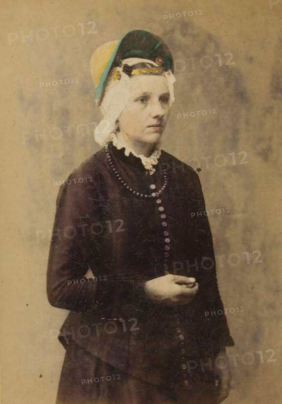 Portrait of an unknown woman with a headdress in the traditional costume of Zaandam, North Holland, La Hollande du Nord. Zaandam, Costumes of the Netherlands, cabinet photograph, inscription: 'Published by A. JAGER, Damrak 81, Amsterdam., Reproduction rights reserved.', retailer's mark: 'Boek-, Papier- en Kunsthandel., Founded 1813, A. VAN HOOGSTRATEN & Zn., Royal Warrant Holders, PLACE 11, - The Hague.', photographer: Andries Jager (possibly), publisher: Andries Jager, Amsterdam, 1860 - 1890, Andries Jager, photographer, publisher, paper, pasteboard, albumen print, height 168 mm x width 108 mm, folk costume, regional costume, historical persons not known by name - BB - woman, A young person shown from head to mid-thigh stands against a plain, textured backdrop. They wear a dark long-sleeved dress with a row of round buttons down the front and a high lace collar. A beaded necklace lies over the bodice. Their hair is covered by a layered headpiece featuring a light cloth, a darker round...
