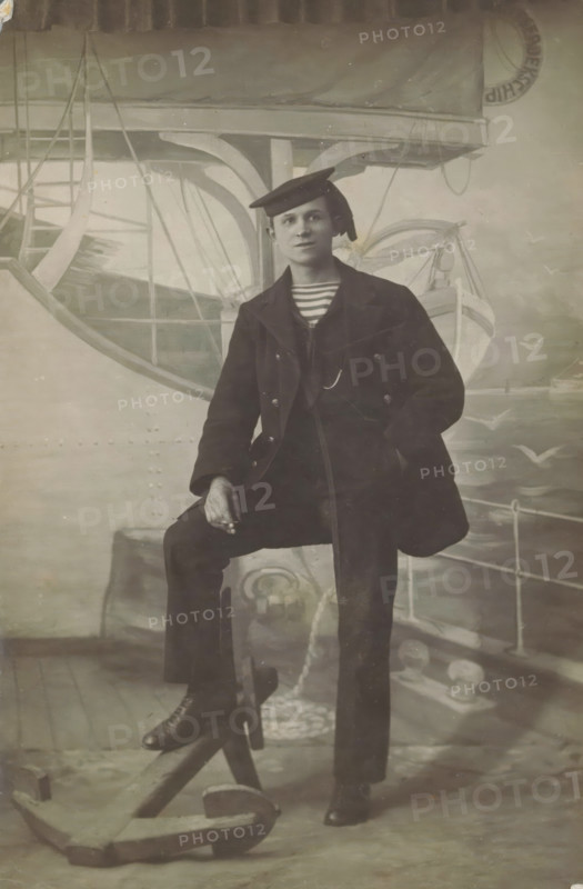 Studio portrait of an unknown man in sailor uniform, photograph, picture postcard, inscription: 'Chef Kreuger Koningstraat 88 Photographer, Den Helder', photographer: Jozeph Kreuger, Den Helder, c. 1915 - c. 1925, paper, gelatin silver print, developing gelatin silver print, height 138 mm x width 90 mm, seaman, studio, A seated person wearing a dark double-breasted coat, striped shirt visible at the collar, dark trousers, and a tilted flat cap sits with one leg raised on a wooden support and one hand resting on the raised knee; the other hand is in a pocket. The backdrop shows painted nautical elements including a lifeboat, rigging, railings, and a large anchor on the floor in front. The image is monochrome with soft lighting and visible surface scratches and creases.