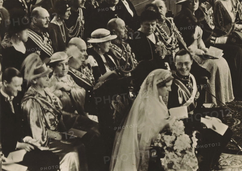 Wedding ceremony of Princess Juliana and Prince Bernhard in the St. Jacob's Church in The Hague on January 7, 1937, photograph, postcard, photographer: Erich Salomon, The Hague, publisher: Het Leven, Weekblad, 1937-01-07, Het Leven, Erich Salomon, photographer, printer (person), publisher, paper, gelatin silver print, height 104 mm x width 146 mm, Person, Juliana of the Netherlands, Bernhard (Prince of the Netherlands), (civil) marriage ceremonies, prince, crown princess, Wedding of Princess Juliana and Prince Bernhard, Grote Kerk (The Hague), A black-and-white photograph showing seated people dressed in formal attire and ceremonial uniforms, many wearing sashes, medals, chains, and hats; a woman in a veil and gown is in the foreground holding flowers, a man beside her in a decorated uniform is smiling, and several other adults in hats, robes, and fur-trimmed coats are seated behind them, some holding booklets.