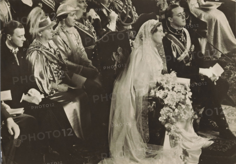 Wedding ceremony of Princess Juliana and Prince Bernhard in the St. Jacob's Church in The Hague on January 7, 1937, photograph, postcard, photographer: Erich Salomon, The Hague, publisher: Het Leven, Weekblad, 1937-01-07, Het Leven, Erich Salomon, photographer, printer (person), publisher, paper, gelatin silver print, height 104 mm x width 146 mm, Person, Juliana of the Netherlands, Bernhard (Prince of the Netherlands), (civil) marriage ceremonies, prince, crown princess, Marriage of Princess Juliana and Prince Bernhard, Flushing, Grote or Sint-Jacobskerk (Vlissingen), A black and white photograph showing a row of seated people dressed in formal attire, including a woman in a bridal gown with a long lace veil draped over her head and shoulders, holding a floral bouquet that cascades forward, and a man next to her in a dark uniform with decorative sashes and gloves holding a folded item. Other seated figures wear hats, ornate robes, and ceremonial garments, and some hold small booklets....