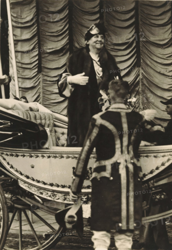 40th anniversary of the reign of HM the Queen, Amsterdam, 5 September 1938, HM the Queen, upon arrival at Haarlemmermeer Station, photograph, postcard, publisher: Have, W. ten, 1938-09-05, photographer, printer (person), paper, gelatin silver print, height 149 mm x width 105 mm, Person, Wilhelmina of the Netherlands, queen, A sepia-toned scene showing a person standing in a decorative carriage wearing a dark fur-trimmed coat and a small hat, smiling and gesturing with one hand while a person with their back to the camera stands in front of the carriage wearing a patterned coat with vertical and diagonal stripes and light-colored footwear; the carriage features tufted upholstery and ornate trim, with visible wooden wheels and a curved side panel, and a draped, scalloped fabric backdrop fills the upper background.