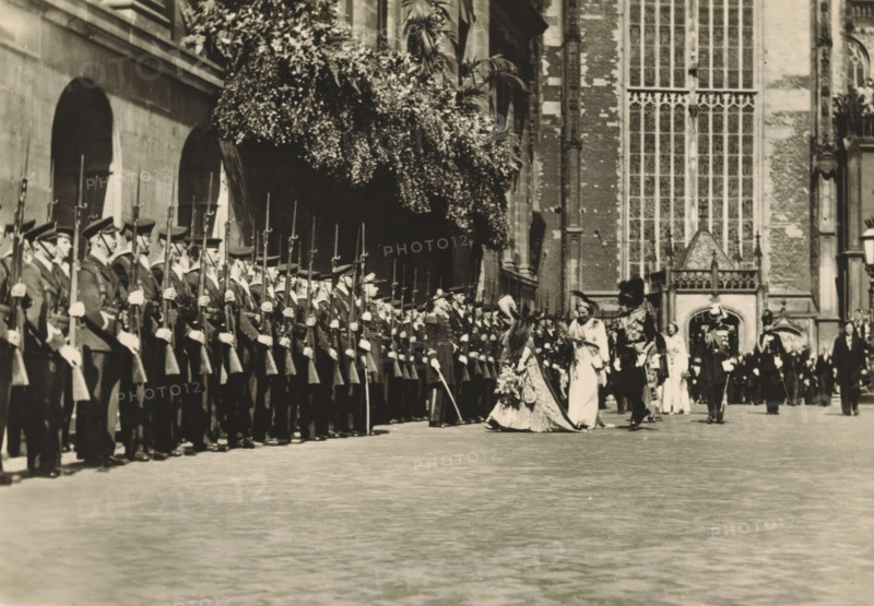 40th anniversary of the reign of HM the Queen, Amsterdam, 6 September 1938. Return from the Nieuwe Kerk, photograph, postcard, publisher: Have, W. ten, 1938-09-06, photographer, printer (person), paper, gelatin silver print, gelatin silver developed, height 105 mm x width 149 mm, Person, Wilhelmina of the Netherlands, queen, Nieuwe Kerk (Amsterdam), A long line of uniformed soldiers stands in formation along the left side of a paved courtyard, each holding a rifle with an upright bayonet and wearing peaked caps and long coats with visible gloves; to the right of them a procession of formally dressed figures moves past, including a central woman in an ornate gown with a long train and floral adornments accompanied by other attendants in light-colored dresses and ceremonial attire, while uniformed officers in decorated uniforms walk nearby; the scene is framed by a large stone building with tall arched windows and a facade of brick and stone, and a dense canopy of foliage overhangs part ...