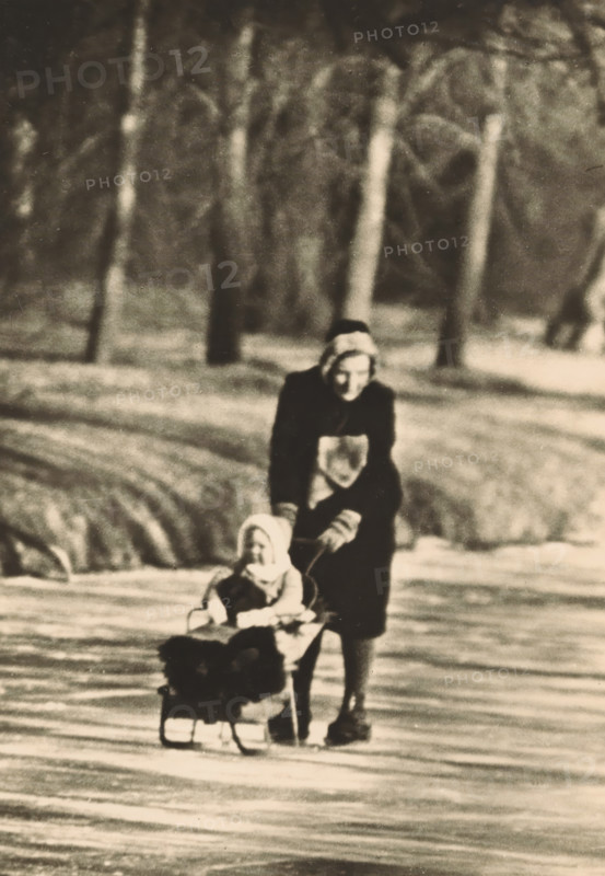 On the ponds of Huis ten Bosch, photograph, postcard, The Hague, publisher: Have, W. ten, Amsterdam, 1940-01-11, photographer, printer (person), paper, gelatin silver print, height 150 mm x width 104 mm, Person, Beatrix (Queen of the Netherlands), Juliana of the Netherlands, mother and daughter(s) (family group), sleigh, sledge (non-sport), princess, crown princess, Huis ten Bosch Palace, A sepia-toned scene showing a person wearing a dark coat and head covering pushing a small child seated in a sled across a snowy surface, with blurred vertical tree trunks and soft-focus snow in the background.