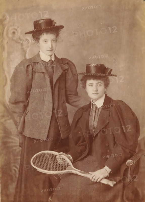 Portrait of two unknown women with a tennis racket, photograph, inscription: 'Bender & Co. 126, George Street, Croydon', number: 'No 1431', inscription: 'Bender & Co. photographic artists, Extra Copies can be had at reduced price.', photographer: Bender & Co., Croydon, c. 1880 - c. 1900, London, paper, albumen print, height 0 mm x width 0 mm, adult woman, tennis, lawn-tennis, A sepia-toned portrait of two young women wearing matching straw hats with ribbon details and high-collared blouses beneath tailored jackets with puffed sleeves, one woman standing with a hand resting behind the other and the other seated in an upholstered chair holding a wooden tennis racquet across her lap; both have short, curled hair peeking from under their hats and neutral facial expressions, with a softly patterned backdrop behind them.