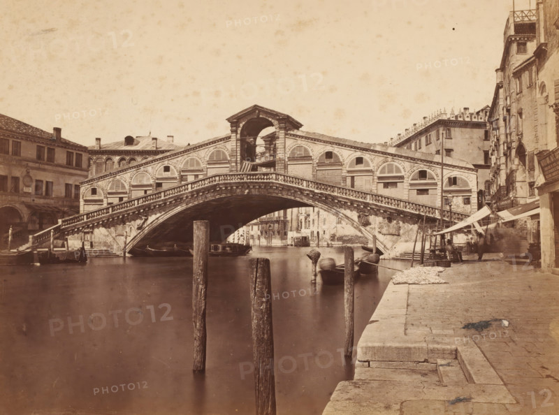 Ponte Rialto, Venice, View of the Rialto Bridge in Venice, Italy, photograph, 1872, photographer, pasteboard, paper, albumen print, albumendruk, height 187 mm x width 256 mm, bridge in city across river, canal, A stone arch bridge spans a calm canal, showing a central rounded opening beneath a balustraded walkway and decorative arched windows along its sides; wooden posts rise from the water in the foreground near small moored boats and gondolas; stone steps and a paved quay run along the right edge with awnings and stalls set up beside adjacent multi-story buildings that have shuttered windows and worn facades; on the left, more buildings with arched ground-level openings and windows frame the canal, and the sky above is pale and textured.