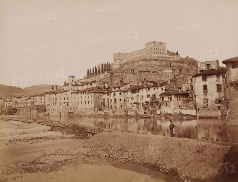 View of Verona and the Adige, Italy, photograph, inscription: 'Verona.', number: '13', number: '68', Verona, 1851 - 1900, photographer, pasteboard, paper, albumen print, albumendruk, height 304 mm x width 405 mm, city-view in general, 'veduta', river, castle, Adige, A riverside scene showing a row of multi-story stone and plaster buildings with tiled roofs lining the water's edge, many with small windows and balconies, some with laundry hanging outside, and small boats moored along the river; behind and above the buildings a steep rocky embankment rises to a large fortified structure with rectangular towers and rows of windows, flanked by a stand of tall narrow trees; in the foreground a gravelly riverbank slopes down to shallow water with a wooden pier or walkway extending along the shore, and a solitary figure standing near the water's edge.