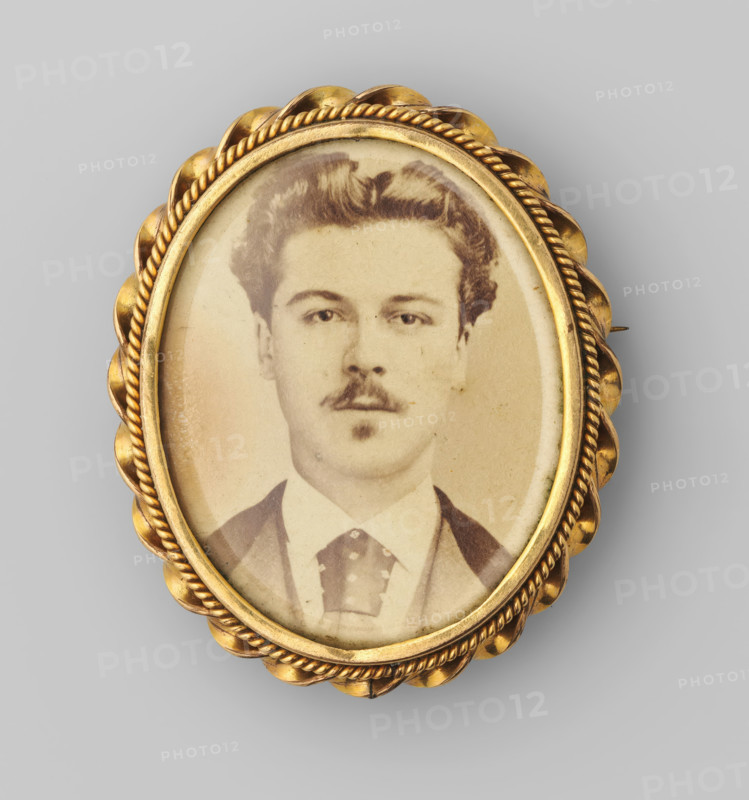 Portrait of an unknown young man with a moustache, photograph, brooch, inscription: 'Paris Montreuil? ff 10 (f 5.50), April 1975', 1851 - 1900, photographer, shell (animal material), biological material, inorganic material, metal, mother of pearl, glass, paper, albumen print, albumen print behind glass in a metal brooch with a mother-of-pearl back, height 46 mm x width 38 mm, historical persons, youth, adolescent, An oval sepia portrait of a man with short wavy hair and a small mustache and goatee is set within an ornate gold frame featuring twisted rope detailing and scalloped outer edges; the man is shown from the chest up wearing a suit jacket, shirt, and patterned necktie, and the framed photograph has a slightly aged, worn surface with small spots and discolorations.