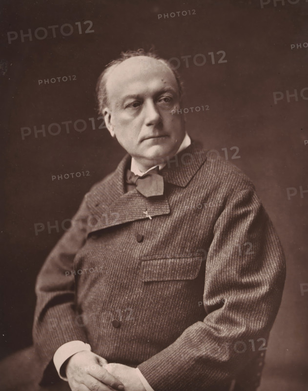 Théodore de Banville, Portrait of Théodore de Banville, page, photomechanical print, inscription: 'Né à Moulins (Allier), le 14 mars 1823.', photographer: Emile Tourtin, cliché maker: Goupil & Cie., c. 1878, paper, Woodburytype, Woodburytypie, height 222 mm x width 178 mm, Person, historical persons, portrait of a writer, A seated adult wearing a textured, striped jacket with a wide collar and a bow tied at the neck is shown from the torso up, hands folded in the lap with visible shirt cuffs, a round forehead with thinning hair, a neutral facial expression and eyes looking slightly to the side, all against a plain dark background.