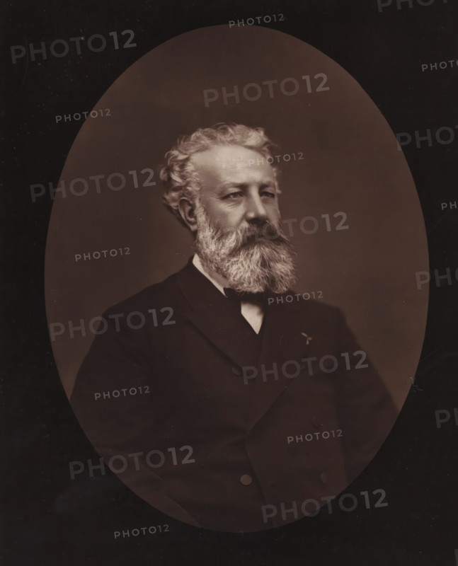 Portrait of Jules Verne, Jules Verne, page, photomechanical print, inscription: 'Galerie Contemporaine, 126, boulevard Magenta. - Paris., Né à Nantes, le 8 février 1828.', photographer: Étienne Carjat, cliché maker: Goupil & Cie., c. 1873 - in or before 1878, paper, Woodburytype, height 231 mm x width 186 mm, Person, historical persons, adult man, An oval-framed sepia portrait of an older man shown from the chest up wearing a dark double-breasted suit, a white shirt collar, and a dark bow tie, with a full, bushy white beard and mustache, wavy white hair swept back, a solemn facial expression, and a softly lit neutral background.