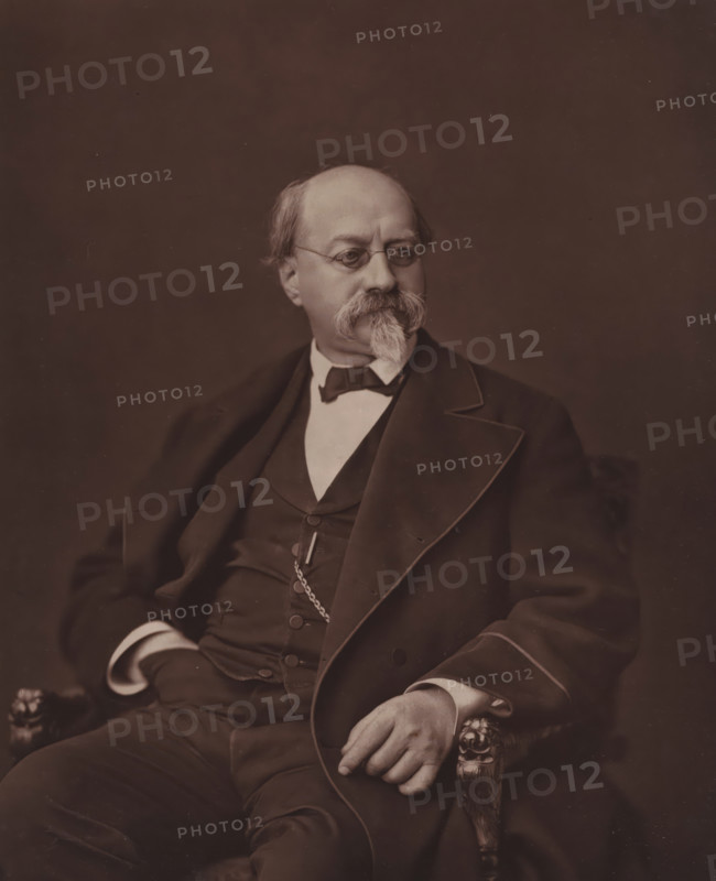 Portrait of Charles-Philippe Robin, Charles-Philippe Robin, page, photomechanical print, inscription: 'Galerie Contemporaine, 126, boul. Magenta. - Paris. (Né a Jasseron (Ain), le 4 juin 1821)', photographer: Ferdinand Mulnier, cliché maker: Goupil & Cie., c. 1874 - c. 1879, paper, Woodburytype, Woodburytypie, height 229 mm x width 186 mm, Person, historical persons, adult man, A seated older man wearing round eyeglasses and a suit with a waistcoat and bow tie is shown from the torso up against a plain dark background, his hair is thinning and he has a large mustache and pointed goatee, one hand rests on an ornately carved armrest while the other is tucked near his waist, and a pocket watch chain is visible across his vest.