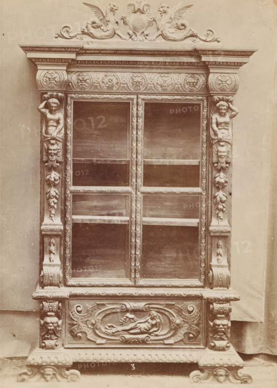 Walnut case, No 3 Mobile Noce, photograph, page, inscription: 'Ia. 320', inscription: 'Alto, 245. Largo, 152. Fondo, 36.', c. 1878 - c. 1885, photographer, paper, albumen print, height 135 mm x width 97 mm, Photo on unnumbered page, ornamental cupboard, An ornate wooden cabinet front with carved decorative elements including symmetrical columns framing a set of glass panels, sculpted human figures integrated into the vertical supports, carved floral and foliate motifs running along the frame, a horizontal frieze decorated with circular carved medallions, a carved crest with winged mythic creatures at the top, rectangular glass panes with visible shelves behind them, a carved lower panel featuring a reclining figure within an oval cartouche, stylized scrollwork and ornamental feet at the base, and a plain backdrop behind the piece.