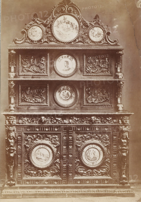 No 5 Buffet, Decorated sideboard, photograph, page, inscription: 'Ies. 350', inscription: 'Alto, 220. Largo, 155. Fondo, 60.', c. 1878 - c. 1885, photographer, paper, albumen print, height 136 mm x width 95 mm, Photo on unnumbered page, ornamental cupboard, A tall ornate wooden cabinet fills the frame, showing multiple carved panels, rounded medallions with painted scenes, and elaborate scrollwork; the top features a decorative crest with three circular painted plaques set within swirling carved foliage, beneath which are two horizontal shelves separated by carved pilasters, each shelf fronted by rectangular panels with raised relief floral and mythical motifs flanking central round painted portraits; lower sections continue the pattern with more carved panels and circular painted medallions encircled by twisted rope borders, all set within a richly carved frame that includes acanthus leaves, human masks, and ornamental borders.