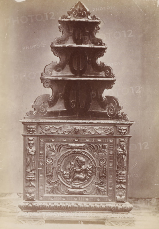 No 14 Cantoniera Noce, Walnut corner cabinet, photograph, page, inscription: 'Alta, 2m. Larga, 1m. Fonda, 68.', inscription: 'Ies 130.', c. 1878 - c. 1885, photographer, paper, albumen print, height 133 mm x width 95 mm, Photo on unnumbered page, ornamental cupboard, A tall ornate carved wooden structure with multiple tiered curved shelves rising to a pointed top, each shelf edged with scrolling motifs and decorative moldings, sits against a plain background. The lower section features a rectangular carved panel with an ornate circular medallion depicting two cherubic figures in bas relief, surrounded by acanthus leaves and scrollwork. Flanking the central panel are vertical pilasters carved with standing draped female figures and additional decorative motifs. The base rests on a molded plinth with carved foliate patterns and a textured surface. The image has a sepia tone and visible surface scratches and marks.