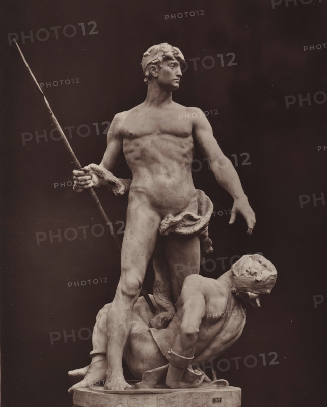 Sculpture 'L'âge de fer' by Alfred-Désiré Lanson, L'âge de fer, page, photomechanical print, photographer: Adolphe Block, cliché maker: Goupil & Cie., after sculpture by Alfred-Désiré Lanson, c. 1879 - in or before 1884, Alfred-Désiré Lanson, after sculpture by, paper, Woodburytype, height 242 mm x width 190 mm, prisoner of war (after the battle), A marble statue of a nude male figure stands with a spear held in one hand and the other arm extended downward, his head turned to the side and hair carved in wavy locks, a draped cloth around his waist, muscular torso and legs visible; at his feet a second partially reclined male figure wears a blindfold and has one arm raised, his body positioned against the standing figure and showing carved details of muscles, hands, and folded cloth; the sculpture rests on a round plinth and is set against a dark plain background.