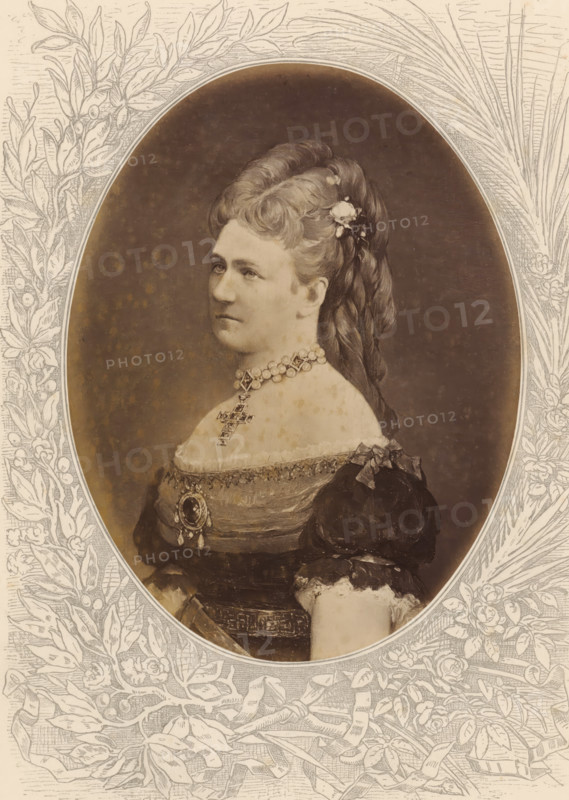 Karoline, Queen of Saxony, Photo reproduction of a painted portrait of Carola, photograph, reproduction, page, after painting by anonymous, c. 1875 - in or before 1880, photographer, paper, albumen print, height 189 mm x width 143 mm, Photo on unnumbered page., Person, Carola (Queen of Saxony), historical persons - BB - woman, queen, empress, etc. (wife of a ruler), A sepia-toned oval portrait of a woman shown in profile from the chest up, set within an ornate floral and leaf engraved border. The woman has hair swept up and braided with a small cluster of flowers pinned near the crown, and she wears a high beaded choker necklace with a pendant and a decorative cross suspended below it. Her dress has puffed sleeves, lace trim at the neckline, and a brooch or medallion at the bodice with additional ribbons and ornamental details. The background inside the oval is plain and darker, while the surrounding border is filled with engraved leaves, flowers, and flowing ribbon motifs.