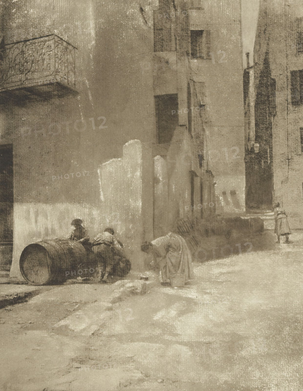 Street corner in Menton, Coin de rue, à Menton, photomechanical print, photographer: Robert Demachy, Menton, cliché maker: Fillon & Heuse, printer (person): Charles Wittmann, Paris, c. 1891 - in or before 1896, printer (person), Provence-Alpes-Côte d'Azur, paper, photoaquatint, height 214 mm x width 166 mm, street, A narrow, muted street scene rendered in sepia tones shows tall, weathered buildings flanking a rough, uneven roadway; a decorative iron balcony projects from the upper left wall, and several recessed windows and doorways punctuate the facades. In the foreground two figures lean over a large wooden barrel lying on its side while another figure bends forward near a small bucket; farther down the street, a solitary figure stands near a corner. The ground is textured with patches of light and shadow, and the overall surface of the image has a grainy, worn appearance.