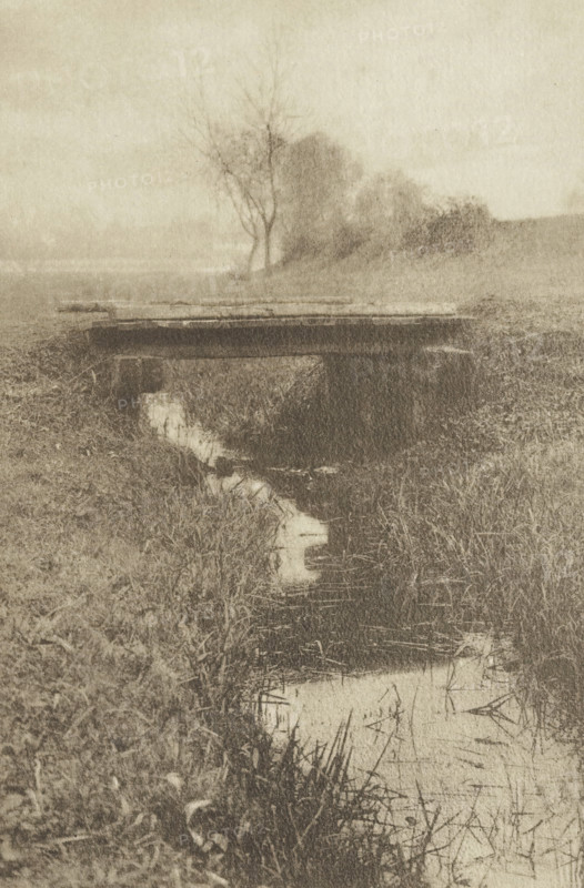 Le Pont, Bridge over a ditch, photomechanical print, photographer: Hugo Henneberg, cliché maker: Fillon & Heuse, printer (person): Charles Wittmann, Paris, c. 1891 - in or before 1896, printer (person), paper, photoaquatint, height 206 mm x width 134 mm, meadow, pasture, water course, bridge, A sepia-toned scene showing a narrow stream bordered by tall grasses and reeds, with a simple wooden plank bridge crossing the stream; the landscape includes a grassy bank, a few leafless and leafed trees in the background, and a misty or hazy sky.
